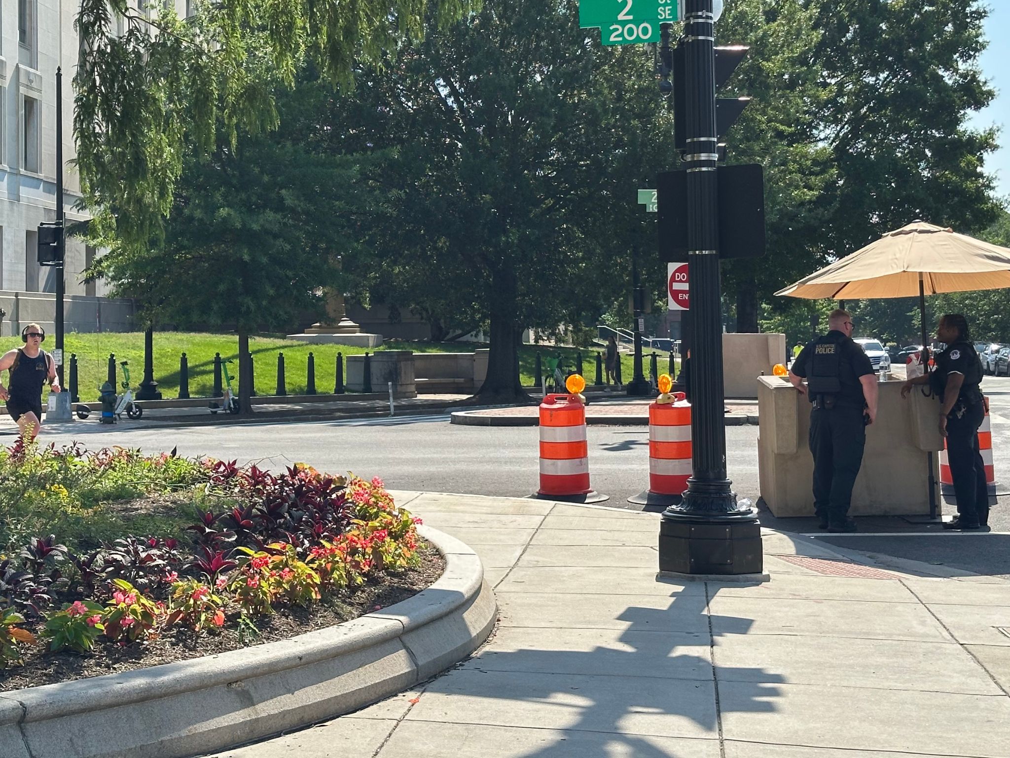View of an intersection in the summertime; two uniformed police officers stand under an umbrella on the right